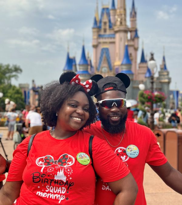Jonise Louis and her husband wear matching red shirts and Mickey Mouse hats as they pose in front of the Disney castle.