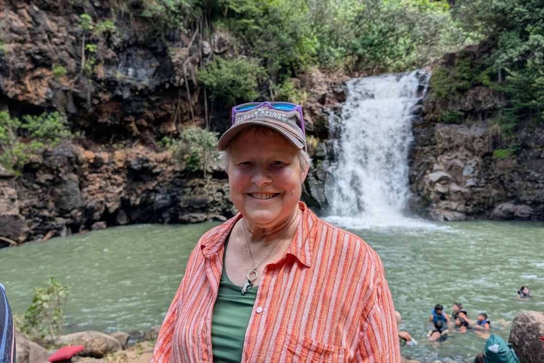 Kim Builta smiles broadly at the beauty of a waterfall and lush, green setting.