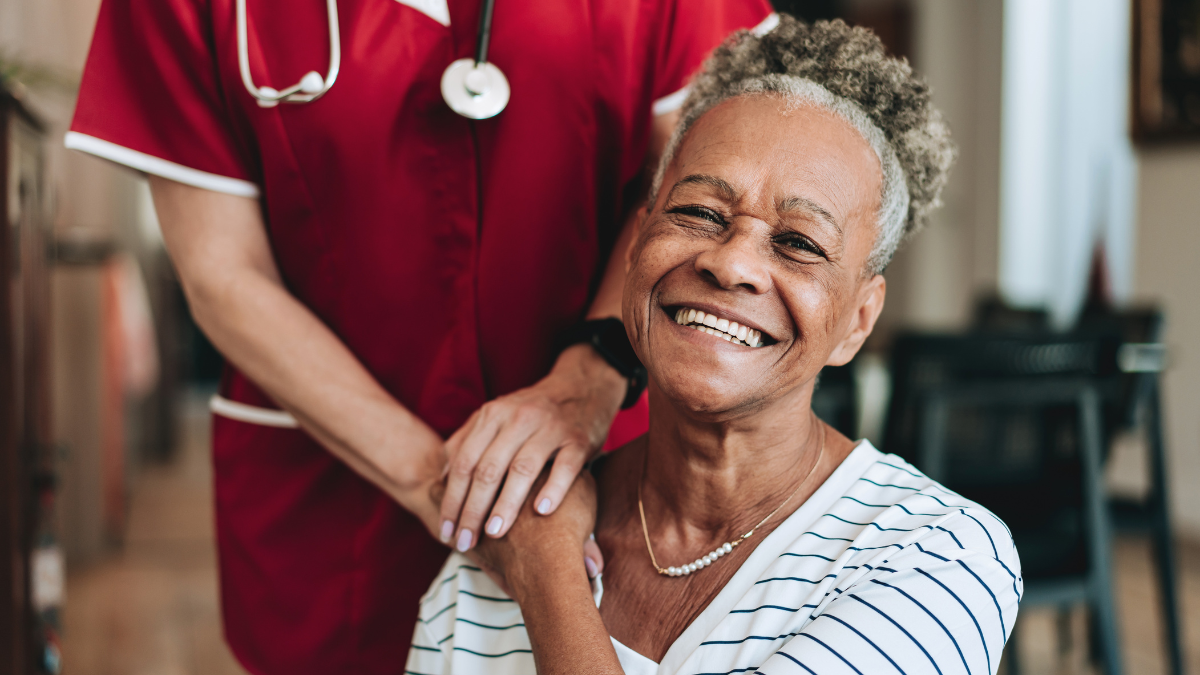A seated older woman is touched on the shoulder by a healthcare worker.