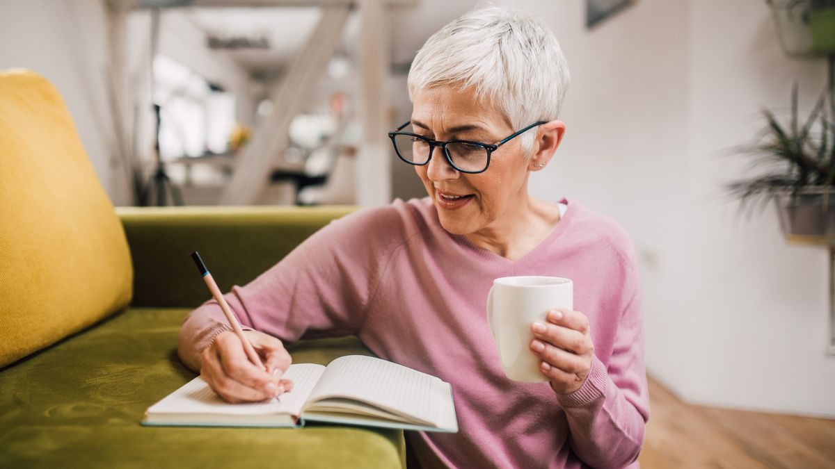 An older white woman journaling in her living room while holding a mug