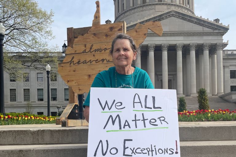 Sherry Lawson sits in front of a state capital building with a placard that says, "We all matter."