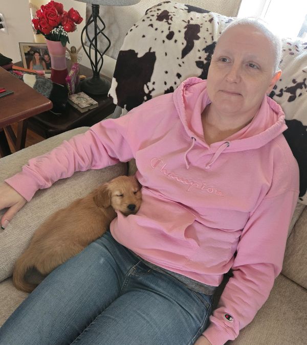 A golden puppy cuddles with Ericka Toth as she sits on the sofa, wearing a pink sweatshirt.