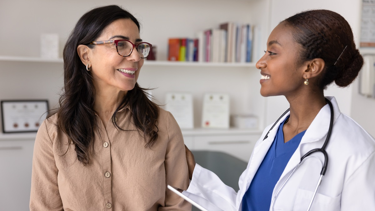 An older woman with glasses and her female, Black doctor smile at each other in a doctor's office.