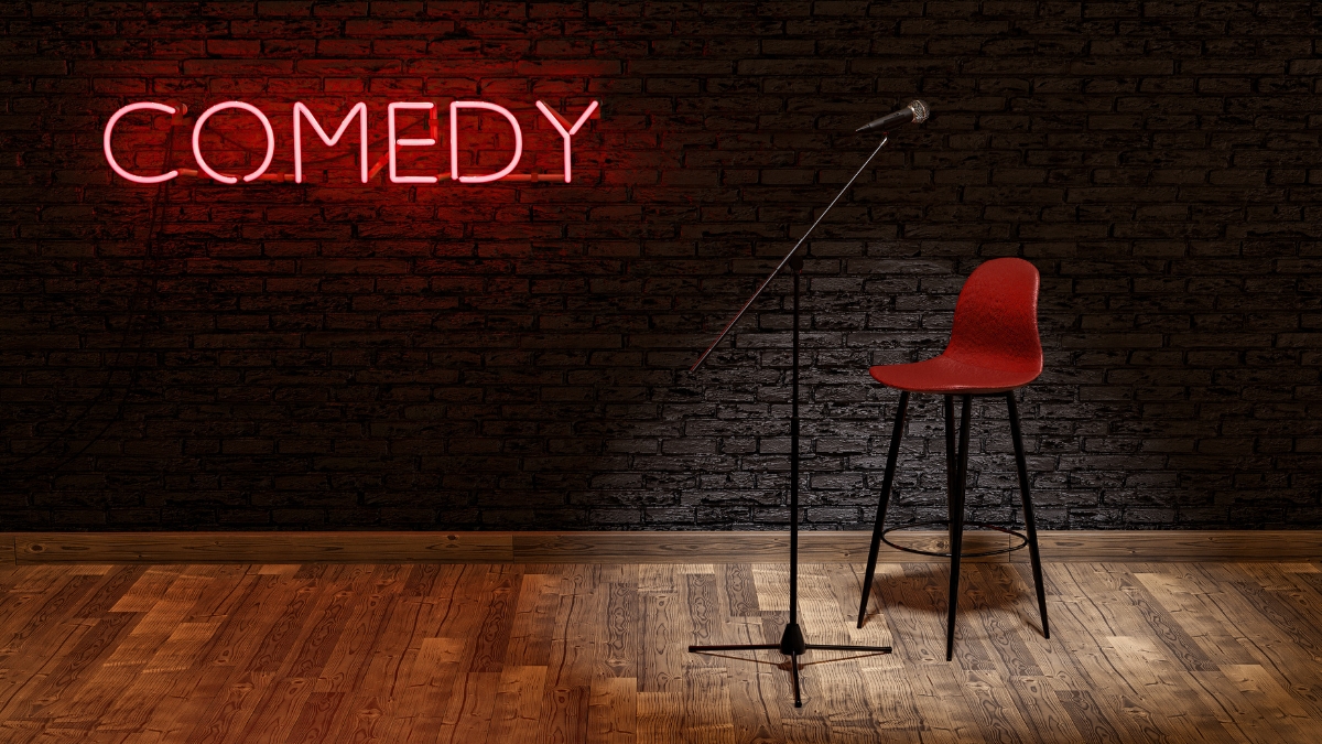 An empty bar stool and a microphone stand on a stage with a neon sign spelling out "comedy" behind.