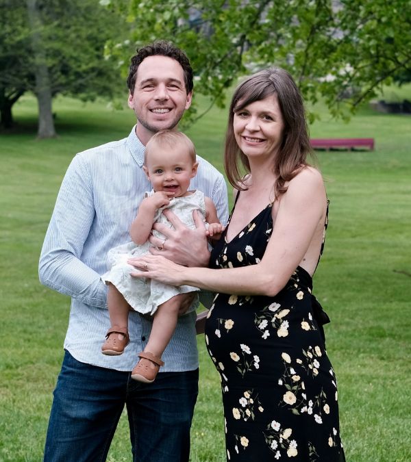 Kim Quick wears a pretty maternity dress as she stands with her husband and infant daughter in a lush, green space.