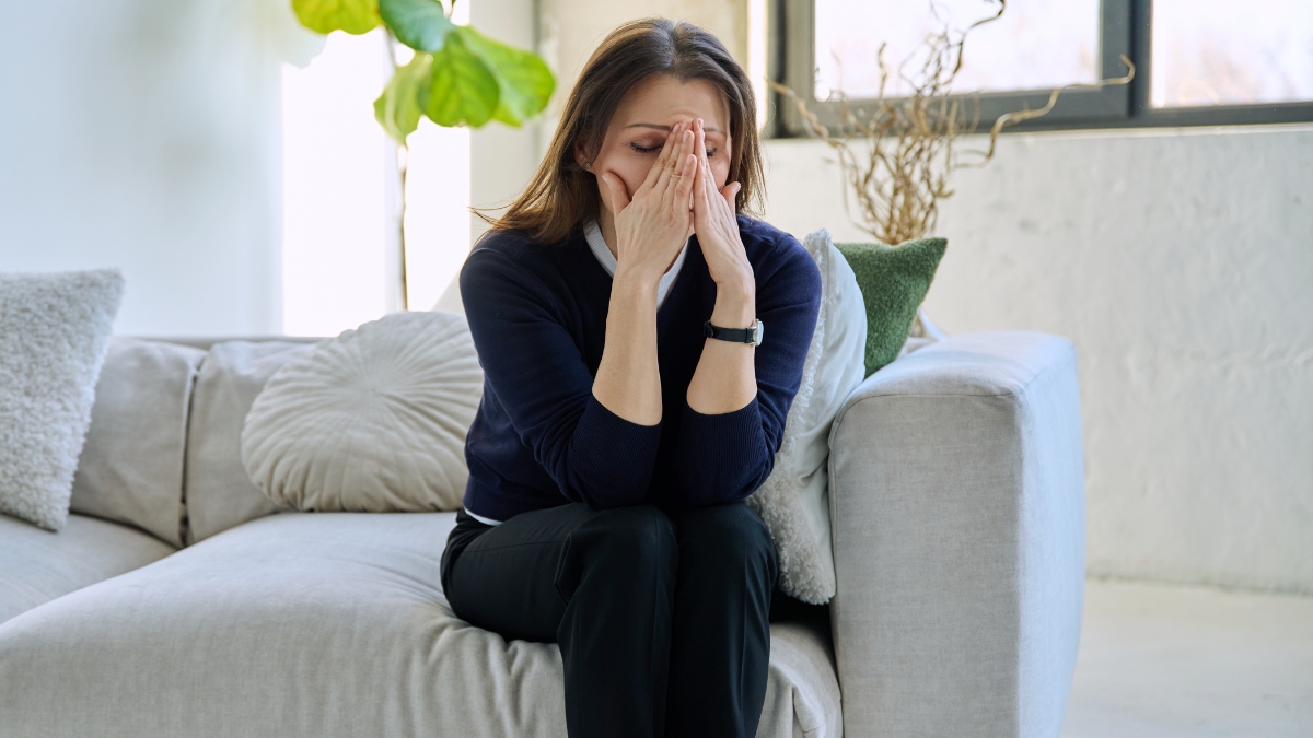 A white woman sits on a sofa with her hands to her face in emotion.
