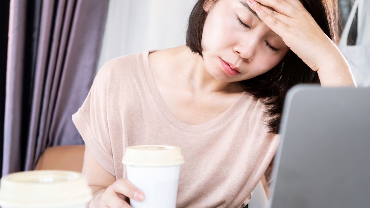 A woman rests her head against her palm as she tries to work, coffee cup in her other hand.