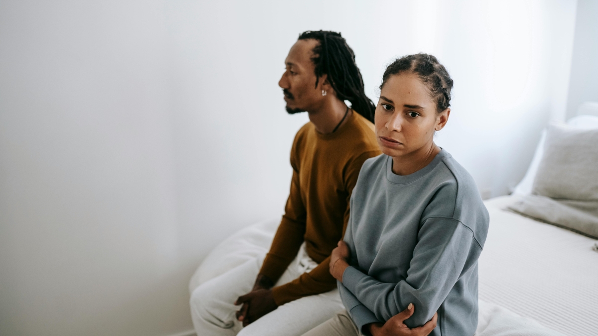 A Black couple sits on the edge of the bed, both looking sadly away from one another.