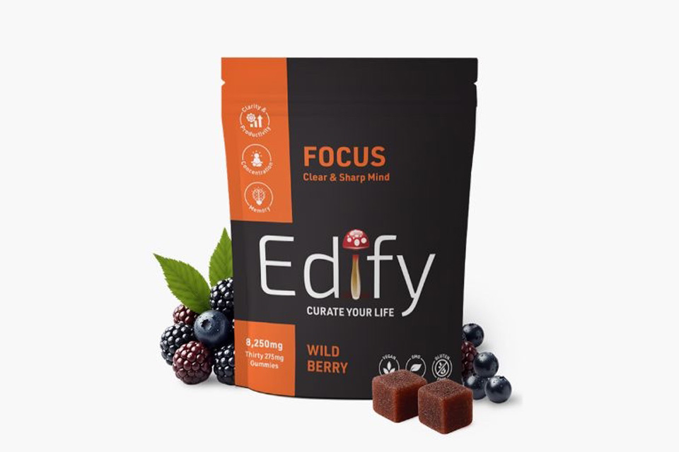 An orange and black bag next to brown cubed gummies and blackberries and blueberries. The bag reads, 