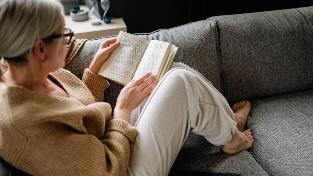 Woman reading a book cozied up on a grey couch.