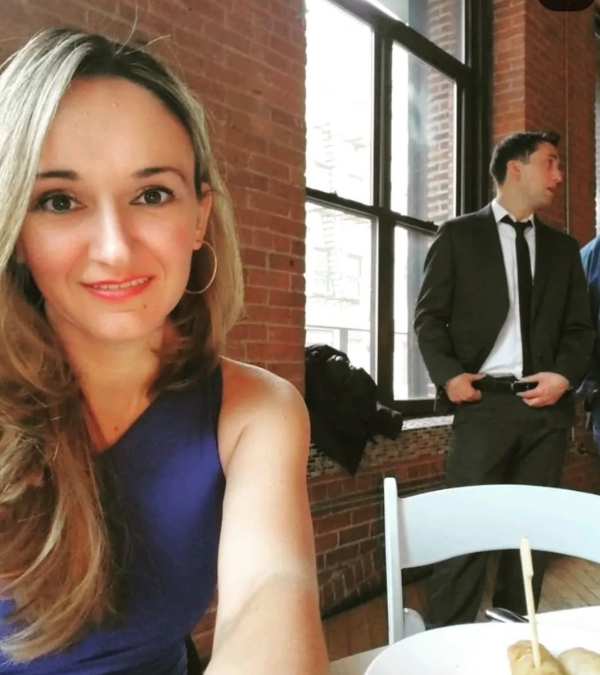 Her hair long and wearing pretty lipstick, Florina Dragovic takes a selfie at a formal event, suit-wearers and rental chairs behind her.
