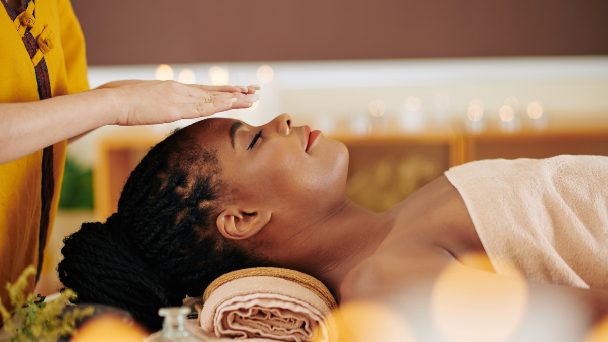 A woman lying with her eyes closed with a reiki practitioner holding their hands above the client's head.