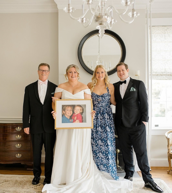 Lilly, Kathryn, Alex, and Mike pose at Kathryn's wedding holding a framed photo of Lauren as a young mom.