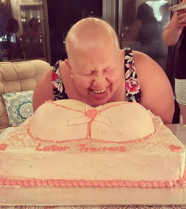 Meg Graves, whose hair has fallen out due to chemotherapy, laughs at a cake decorated with two breasts, titled "Later Traitors."